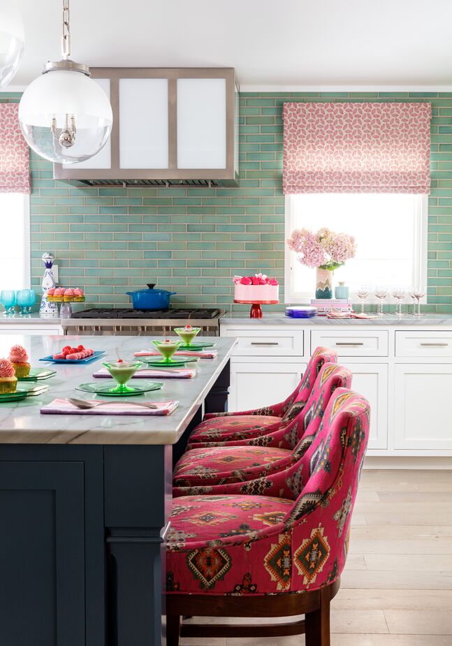 White kitchen cabinets are anything but boring when complemented by a soft green backsplash and stools with vibrant pink upholstery. The island was painted blue to tie in with the palette of the adjacent dining room.
